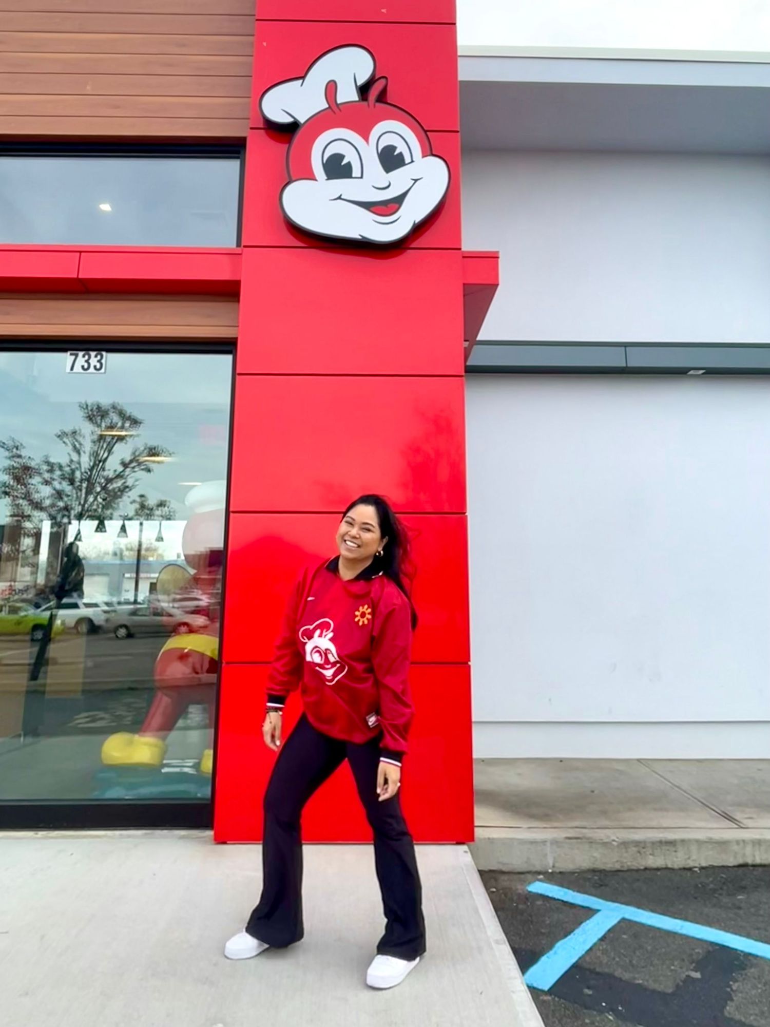 Person in red sweater poses cheerfully by cartoon mascot sign on bright red building exterior.