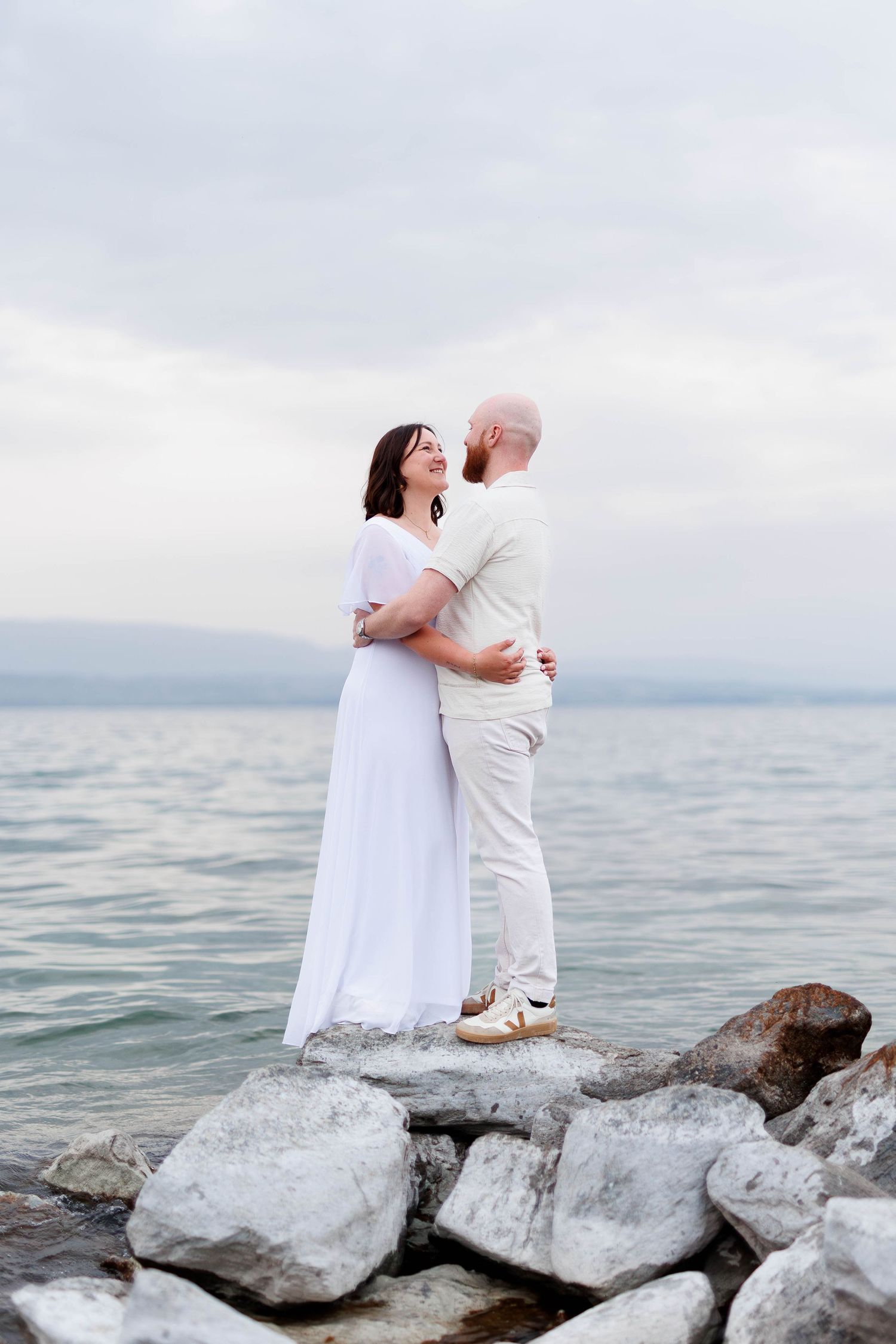 Couple embracing on rocky shoreline, woman in white dress, man in beige outfit, with calm sea and mountains in background.