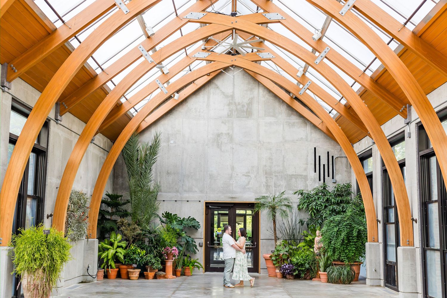 Couple embraces under stunning wooden arched ceiling with skylight surrounded by lush tropical plants in elegant greenhouse venue.