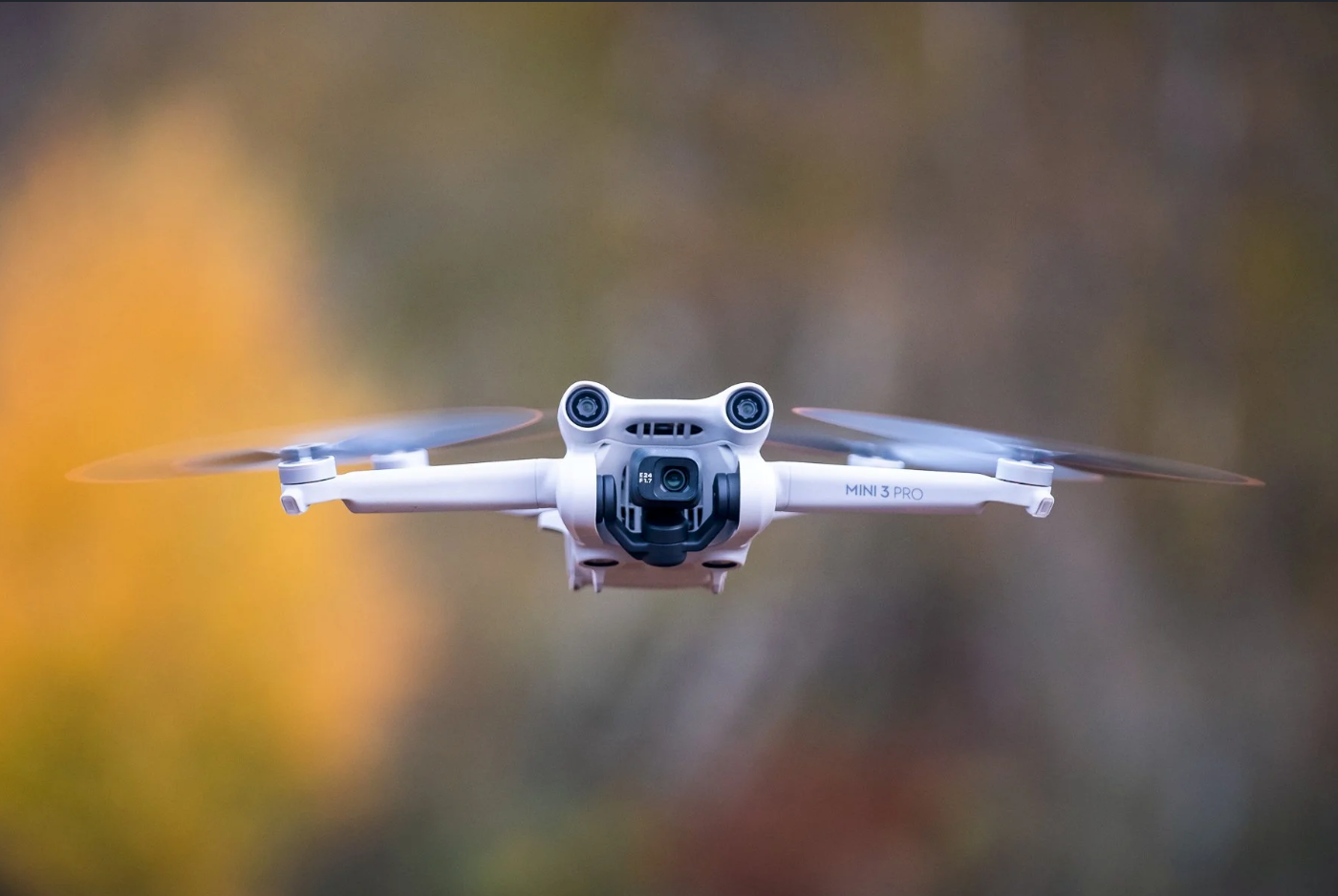 A DJI Mini drone with camera hovering in mid-air against a blurred autumn foliage background.