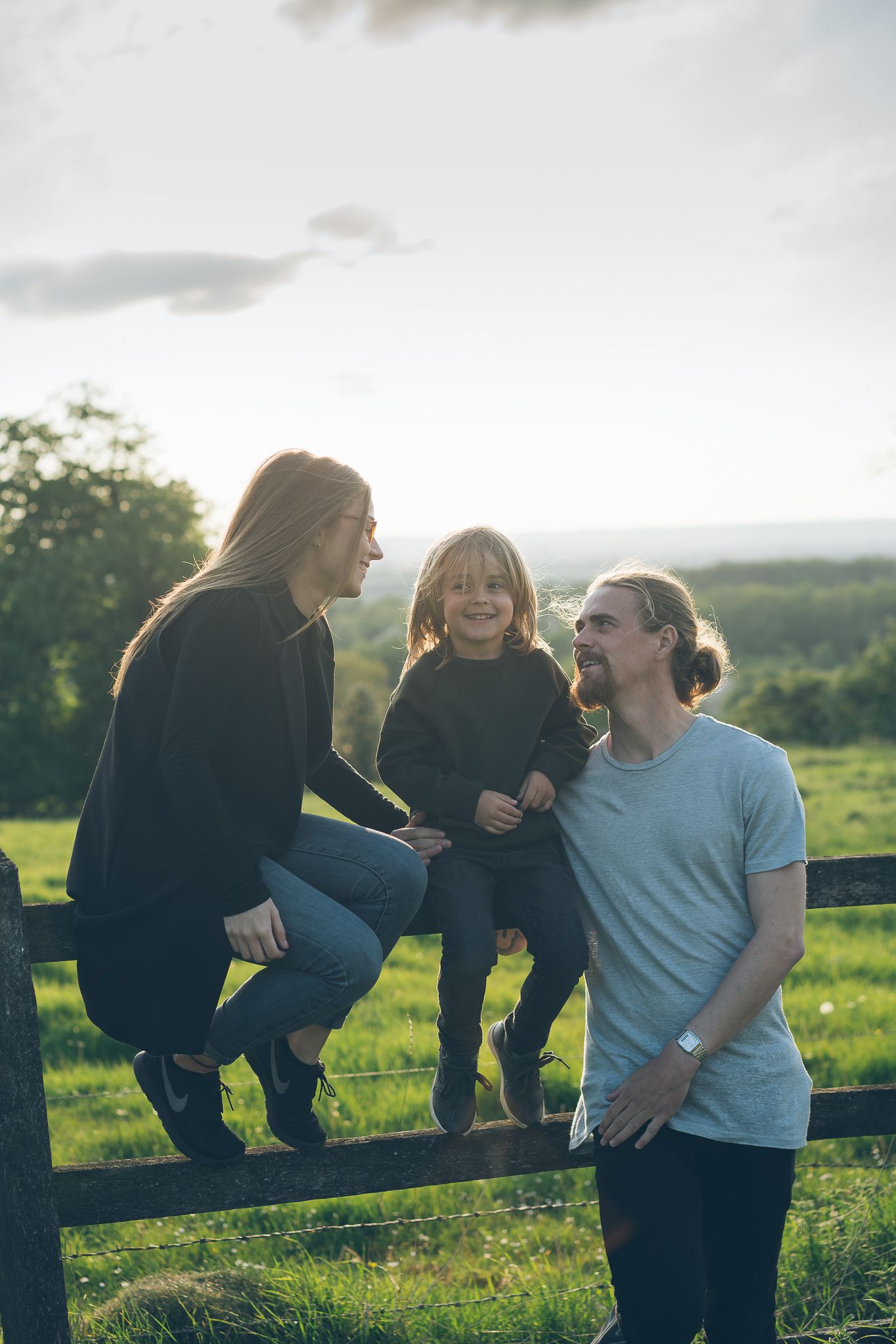 A happy family with a young child sitting on a wooden fence in a lush green field at golden hour.