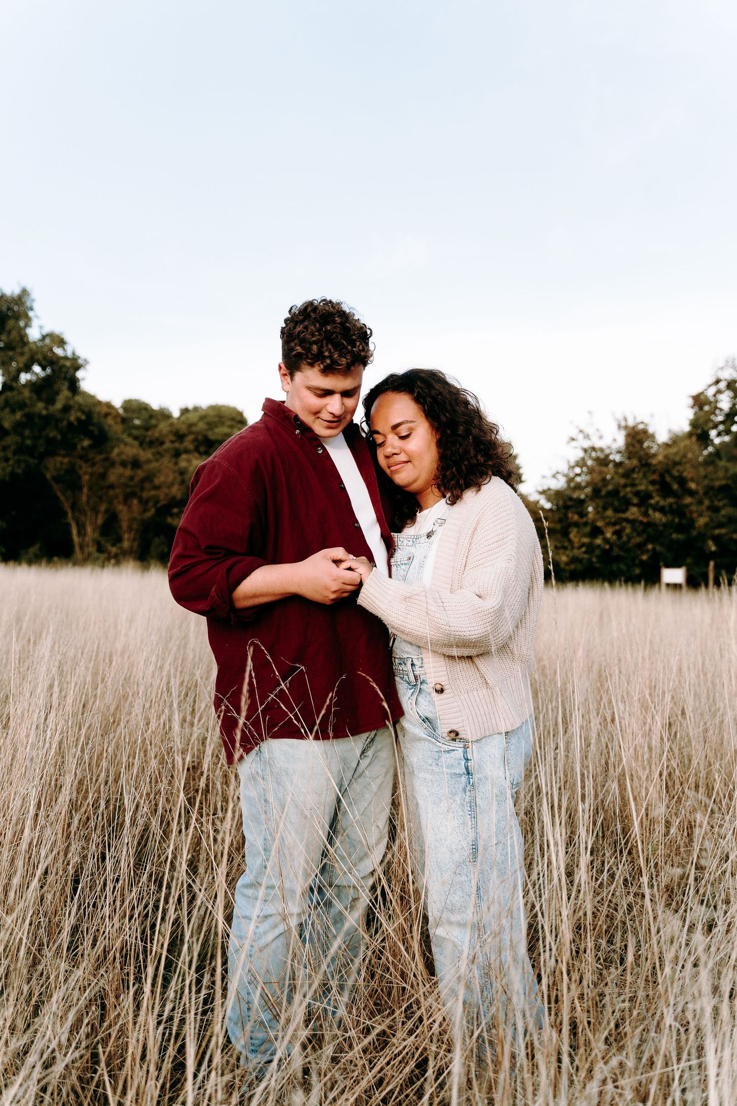 A romantic couple embraces in a golden field of tall grass during sunset wearing matching denim and burgundy outfits.