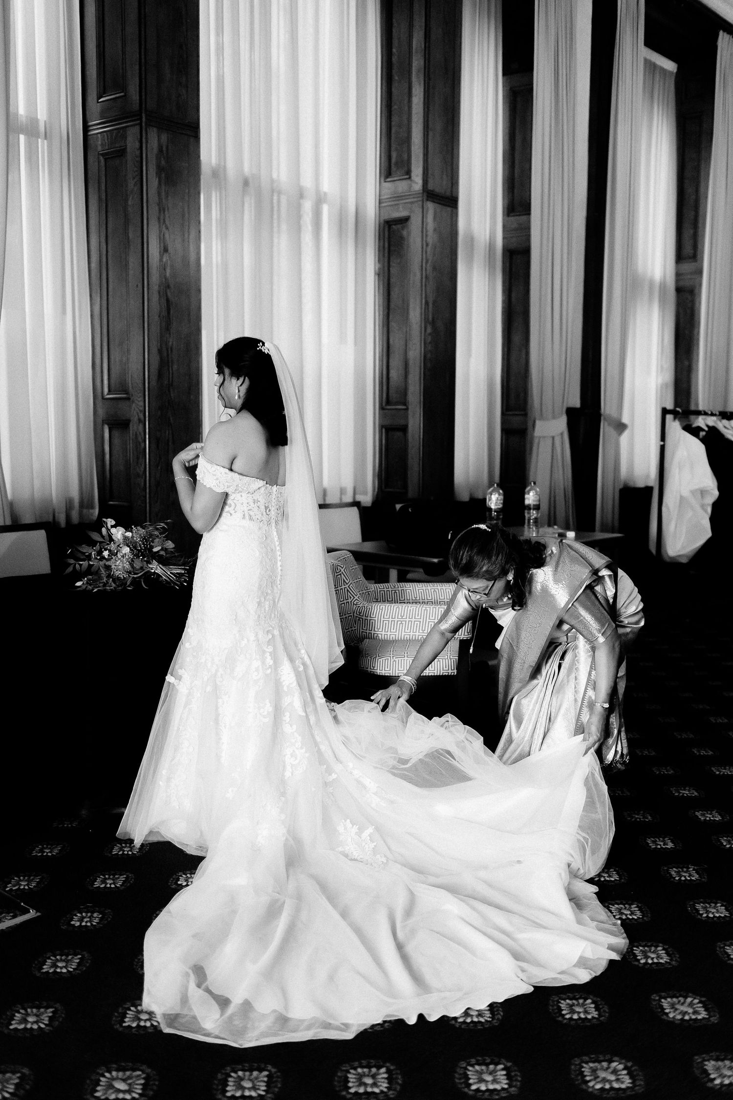 A black and white photograph showing someone in a flowing white wedding dress with a long train being adjusted.