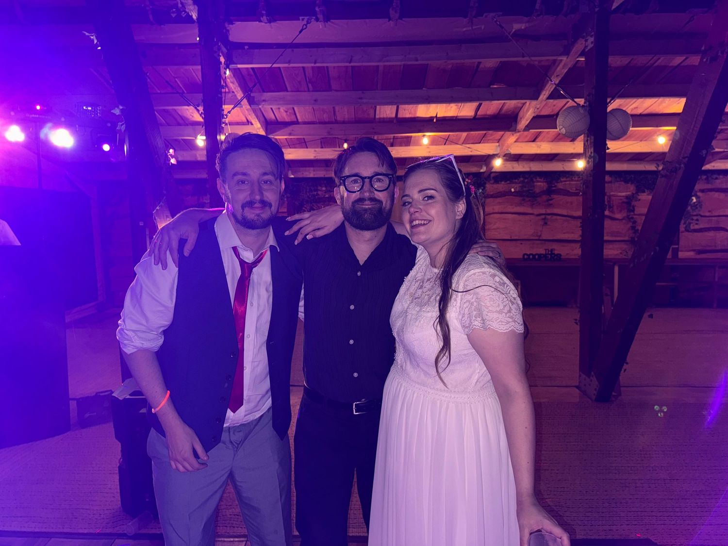 Three people posing together at a wedding reception in a rustic barn venue with purple lighting.