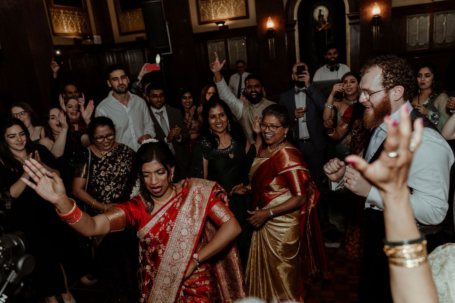 Bride in red saree dancing joyfully with guests at a vibrant Indian wedding reception celebration. London