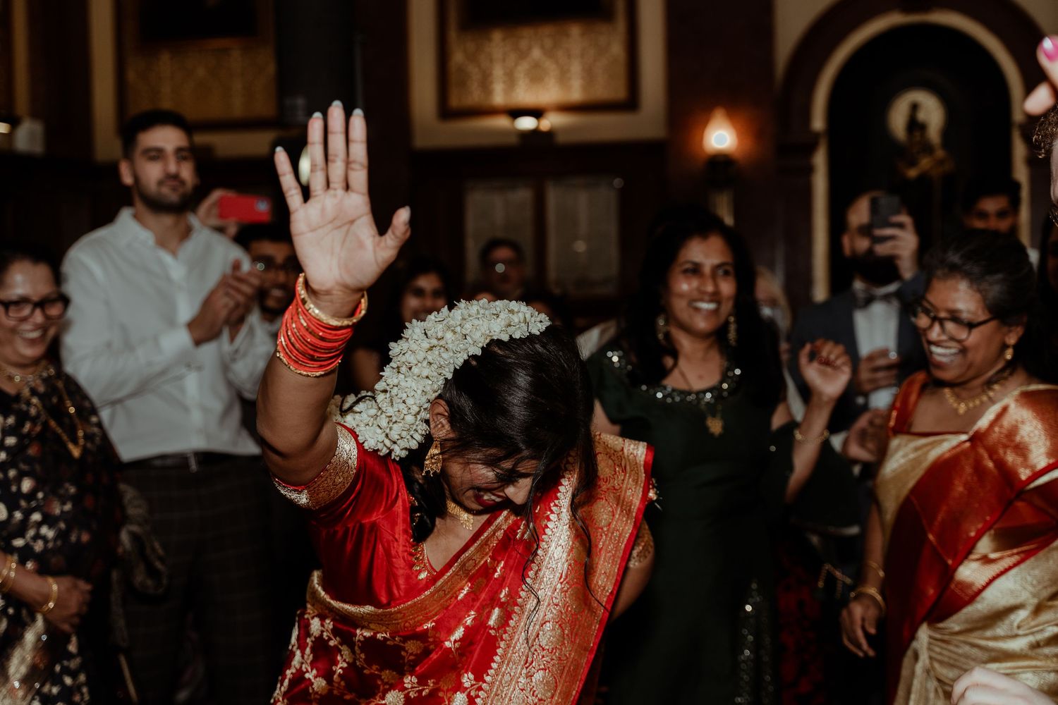 A bride in a red saree with floral hair accessory raises her hand joyfully during a traditional Indian wedding celebration.
