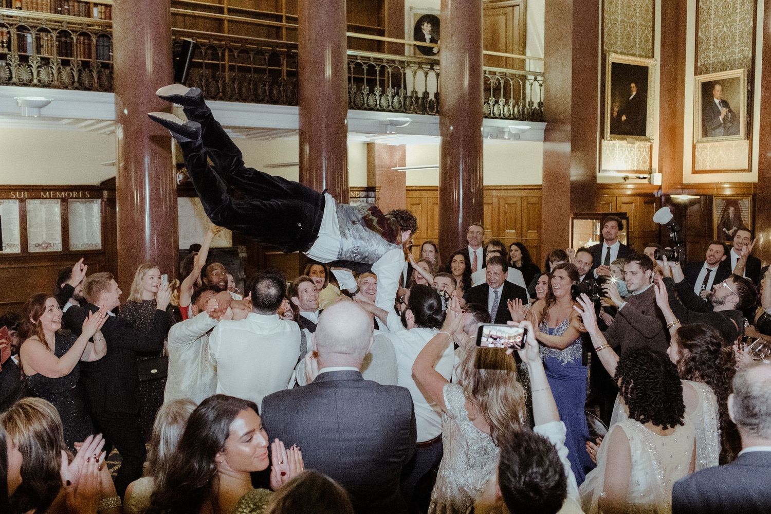 Guests lift a man into the air during a lively wedding reception celebration inside an elegant venue.