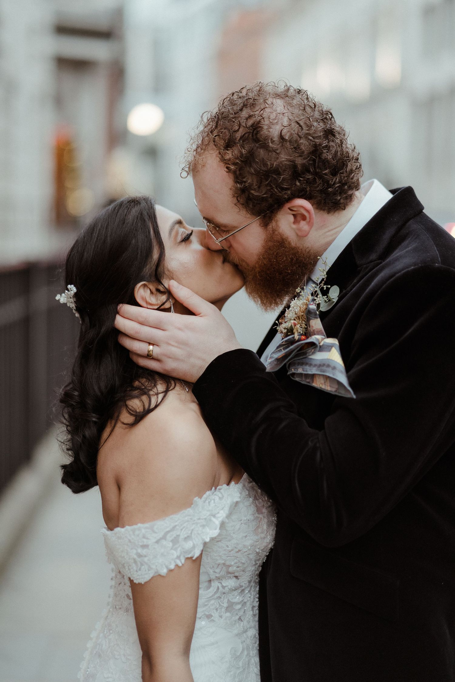 A bearded groom in a black velvet suit kisses his bride in a white lace off-shoulder gown outdoors.