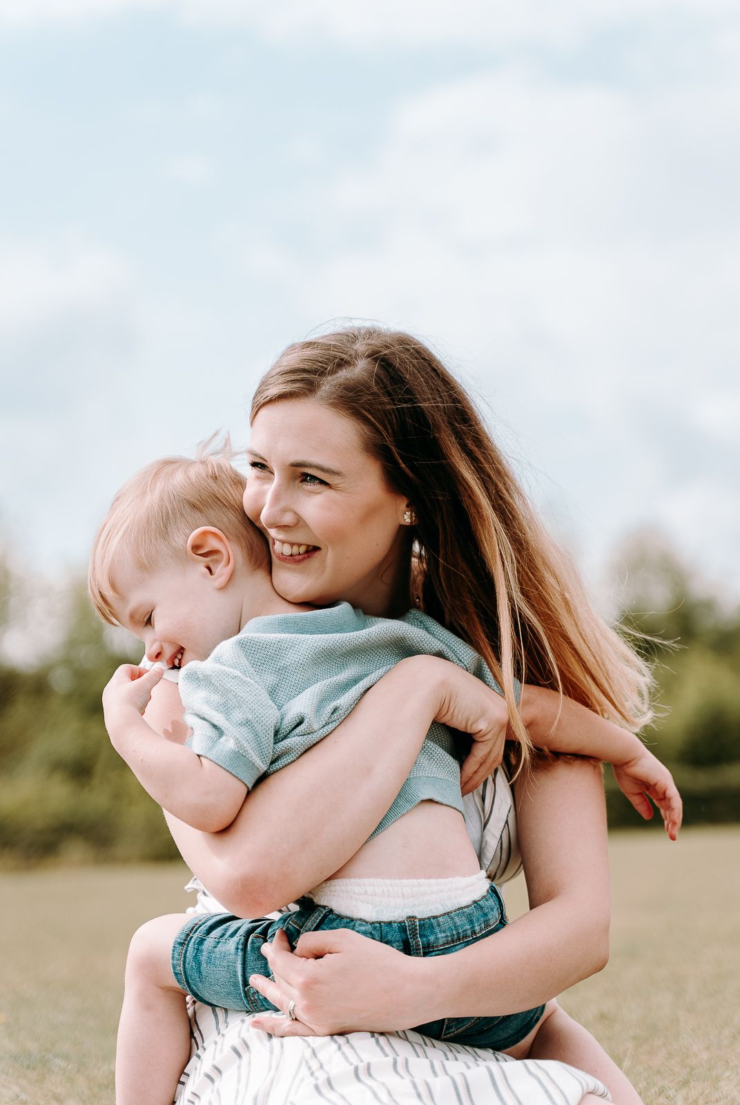 A smiling young woman holds a happy toddler in a sunny green field under a partly cloudy sky.