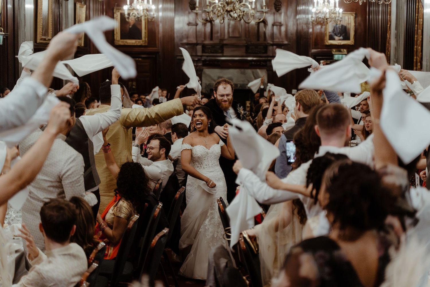Joyful wedding reception with bride and groom surrounded by cheering guests waving white napkins in elegant ballroom.