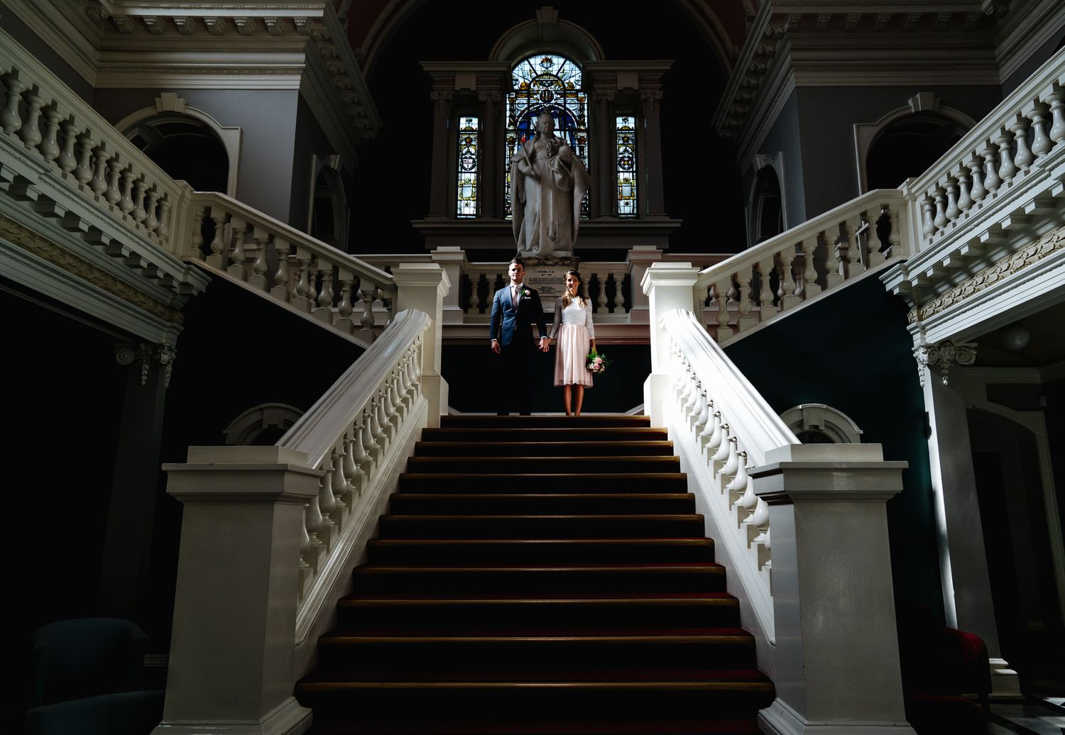 Couple stands on grand staircase in elegant historic building with stained glass windows. Woolwich Town Hall, London