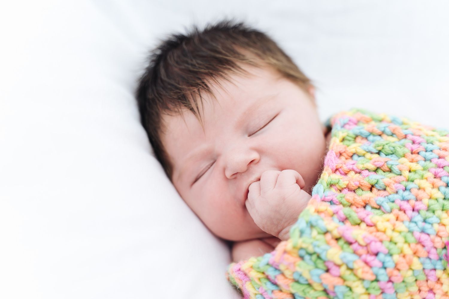 Peaceful newborn baby sleeping wrapped in a colorful pastel knit blanket with hand near face on white background.