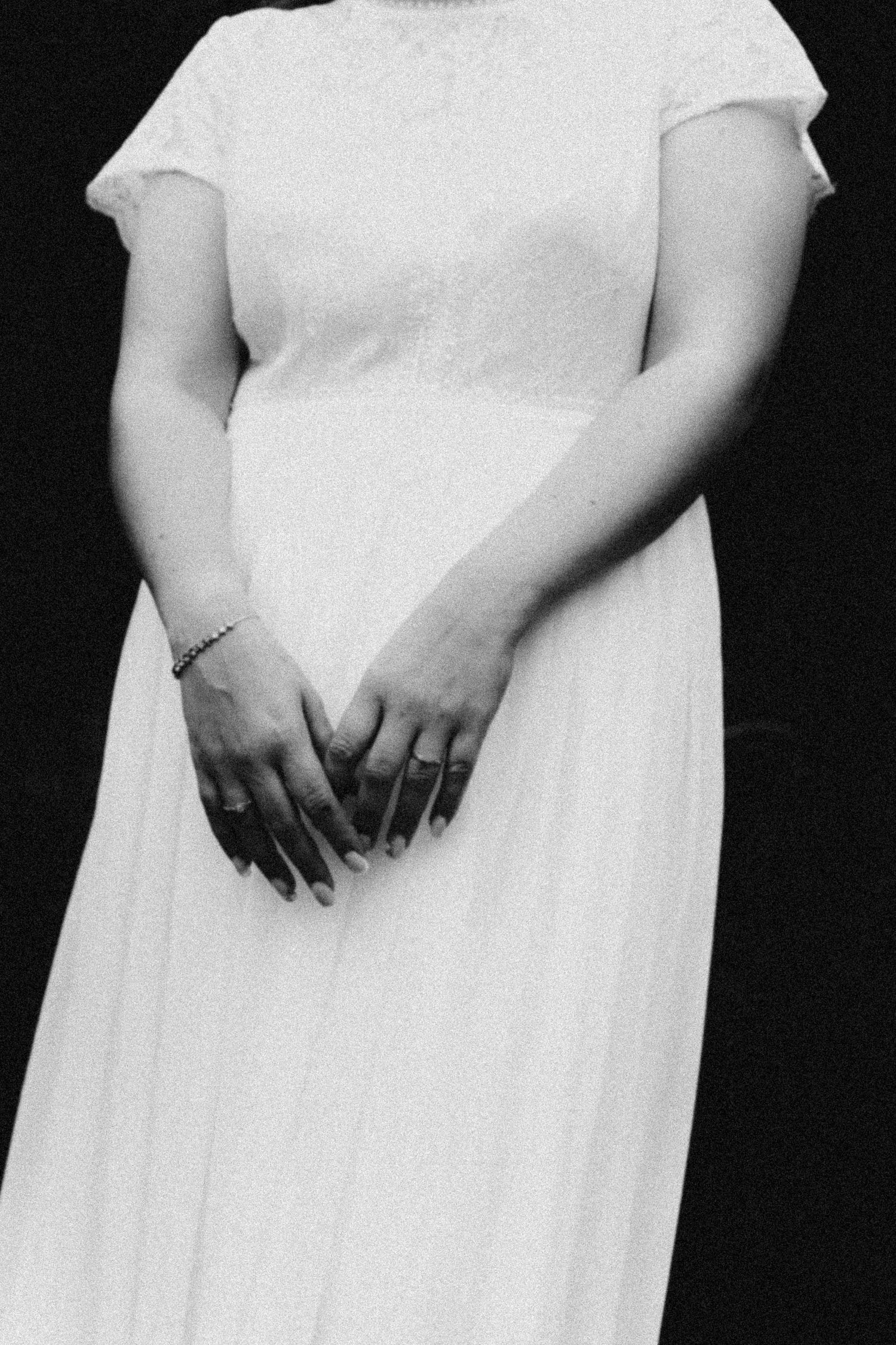 Black and white photograph showing hands clasped in front of a flowing white dress with short sleeves.