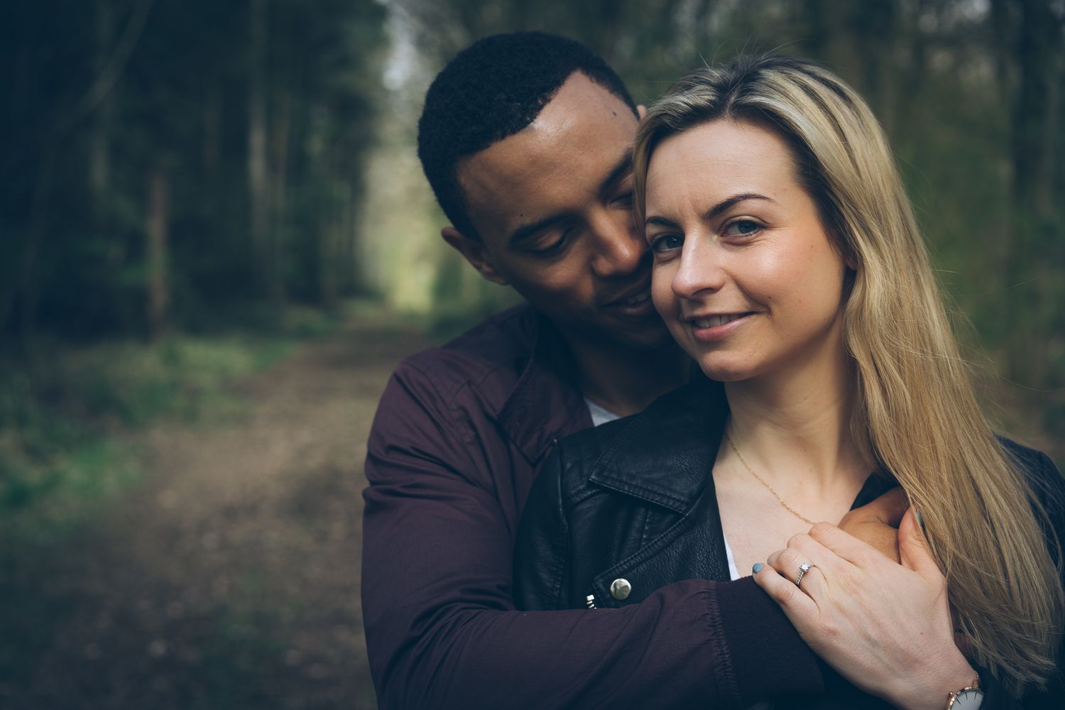 A loving interracial couple embraces in a forest, the man tenderly kissing the smiling woman's cheek.
