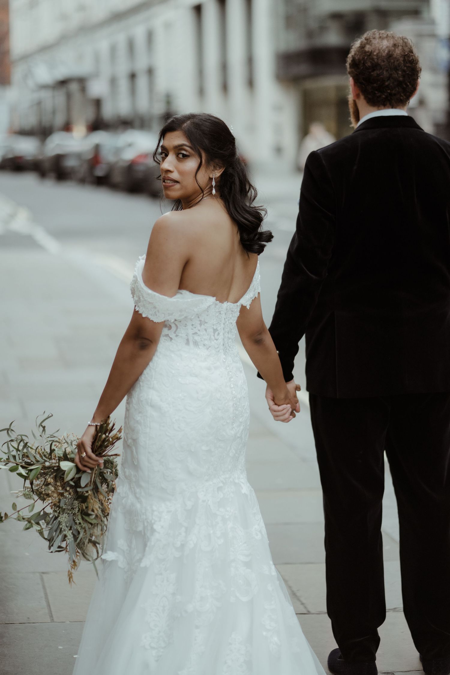 Bride in off-shoulder lace mermaid gown holds bouquet, walking hand-in-hand with groom in black suit on London city street.