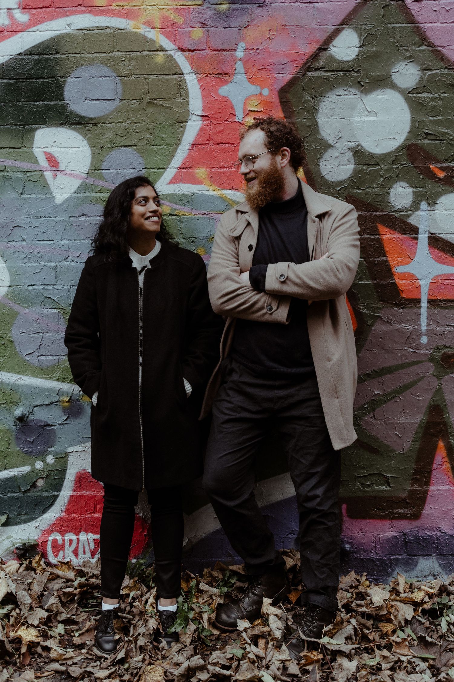 A couple shares a playful moment against a colorful graffiti wall covered with autumn leaves.