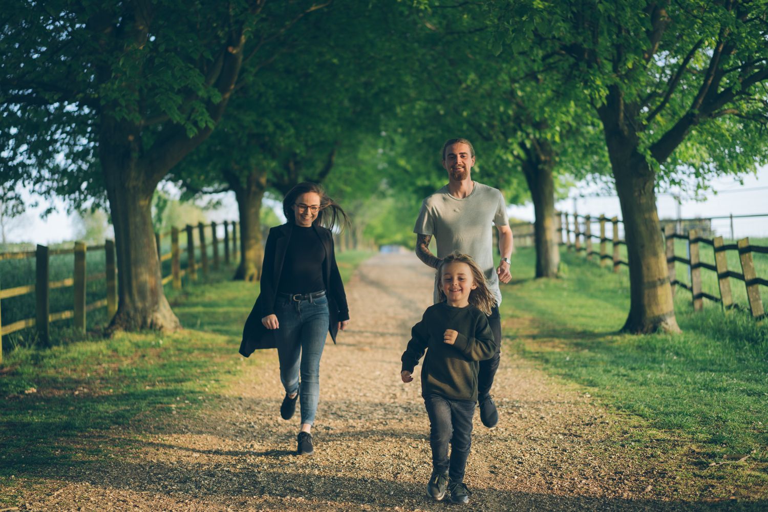 Family of three running joyfully down a tree-lined country path on a sunny day.