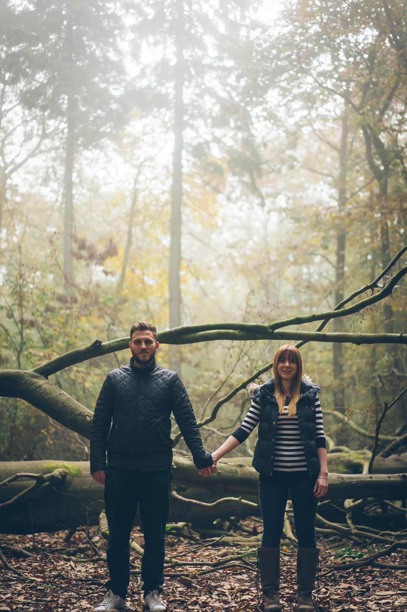Couple holding hands in a misty autumn forest with fallen trees and golden foliage in the background.