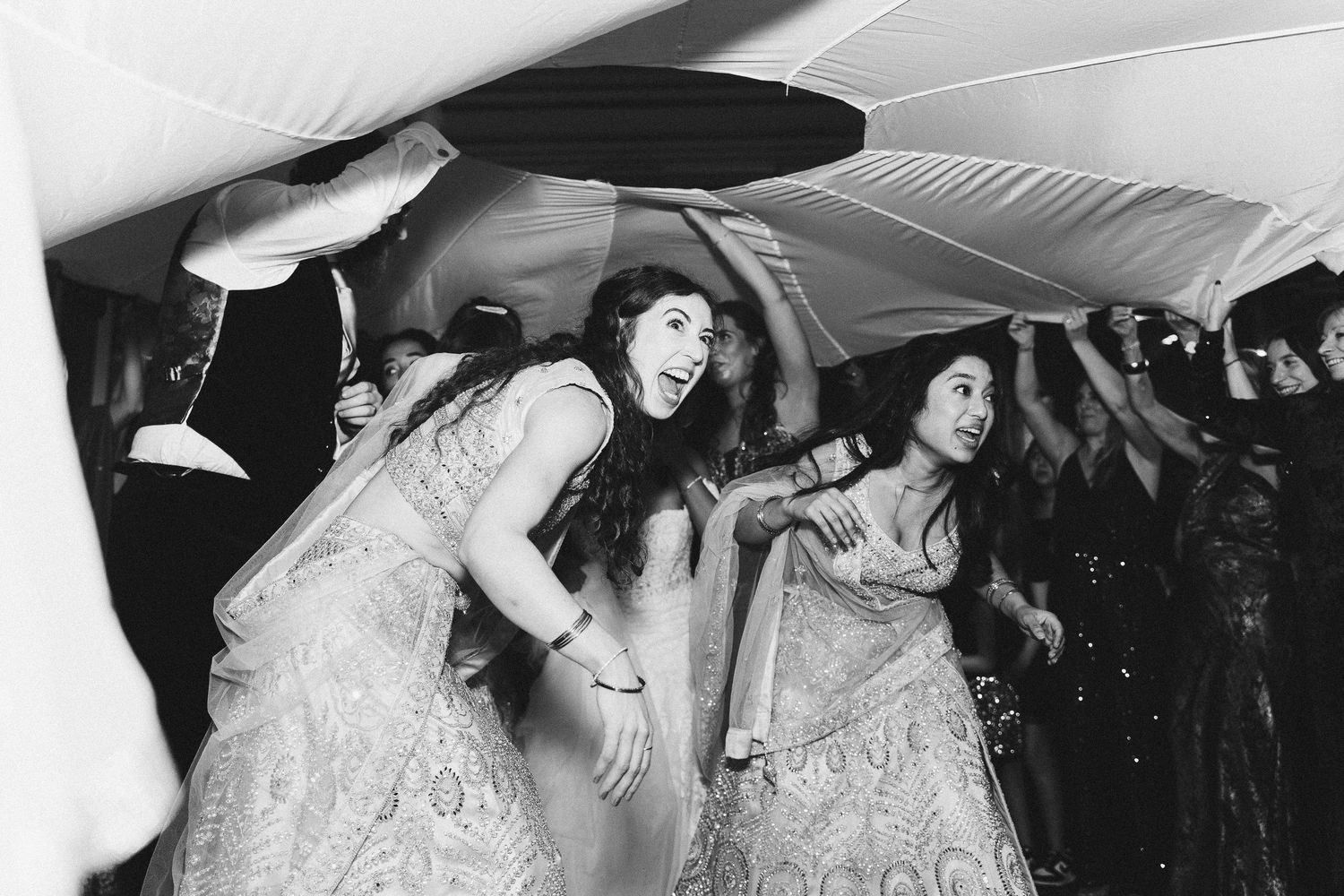 Women in traditional Indian attire dancing joyfully at a wedding celebration, captured in black and white.