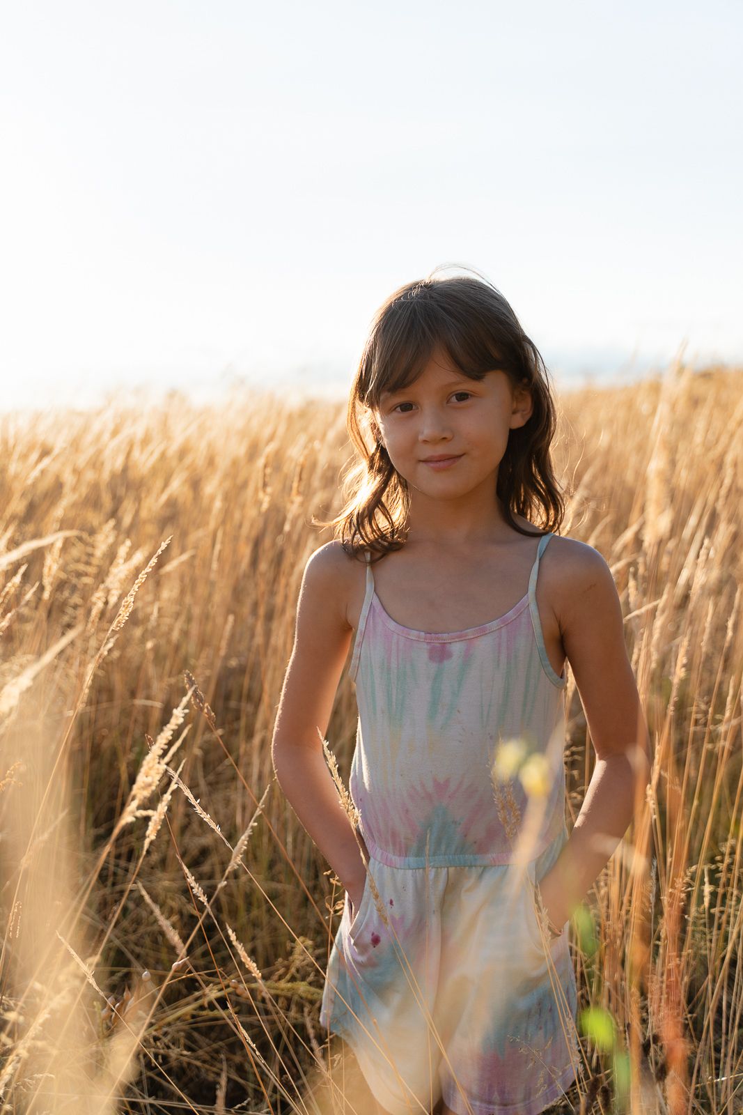Young girl in a floral romper standing in a golden wheat field during sunset.