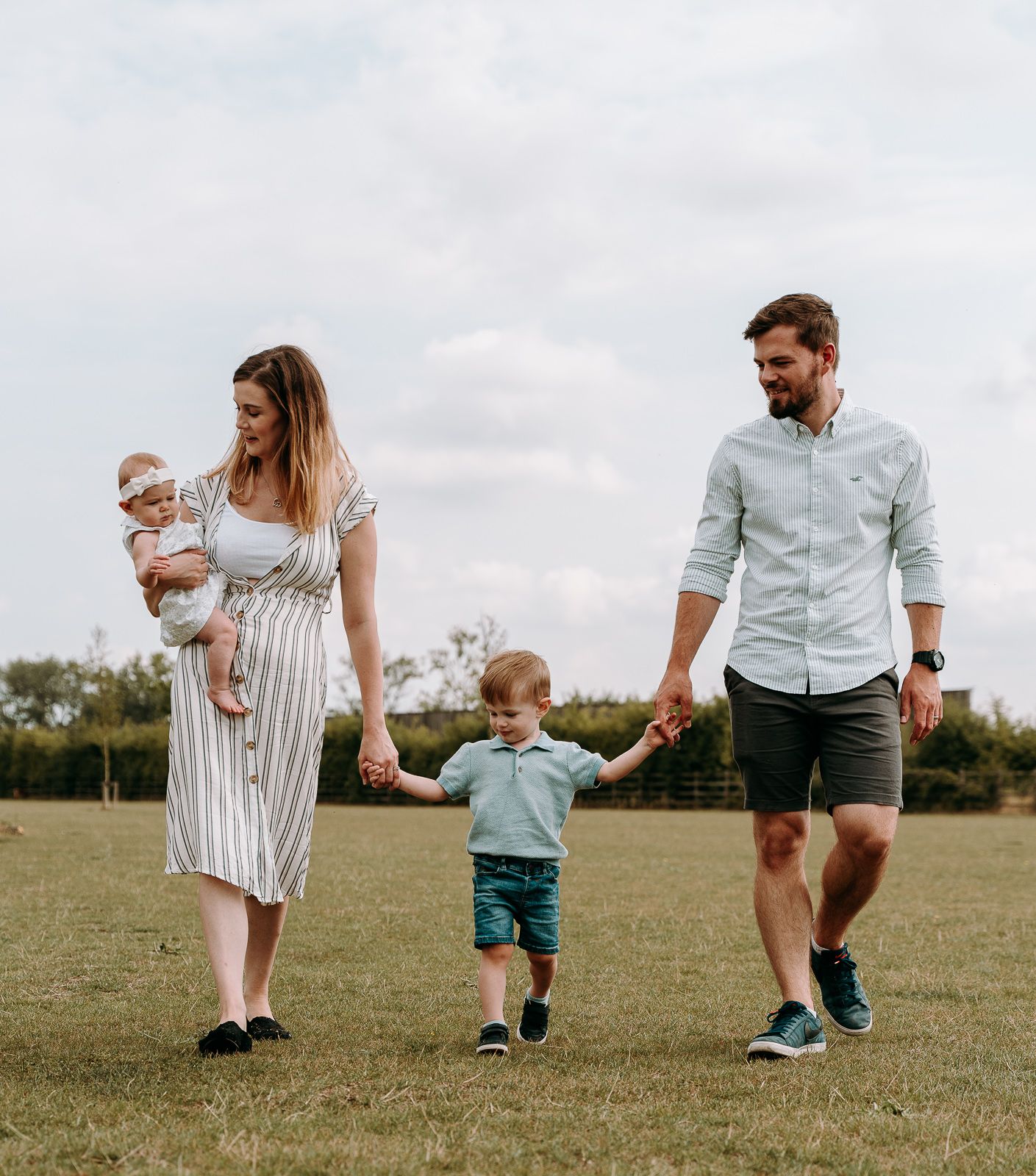 A happy family of four walks through a green field, with mom holding a baby and dad holding their toddler's hand.