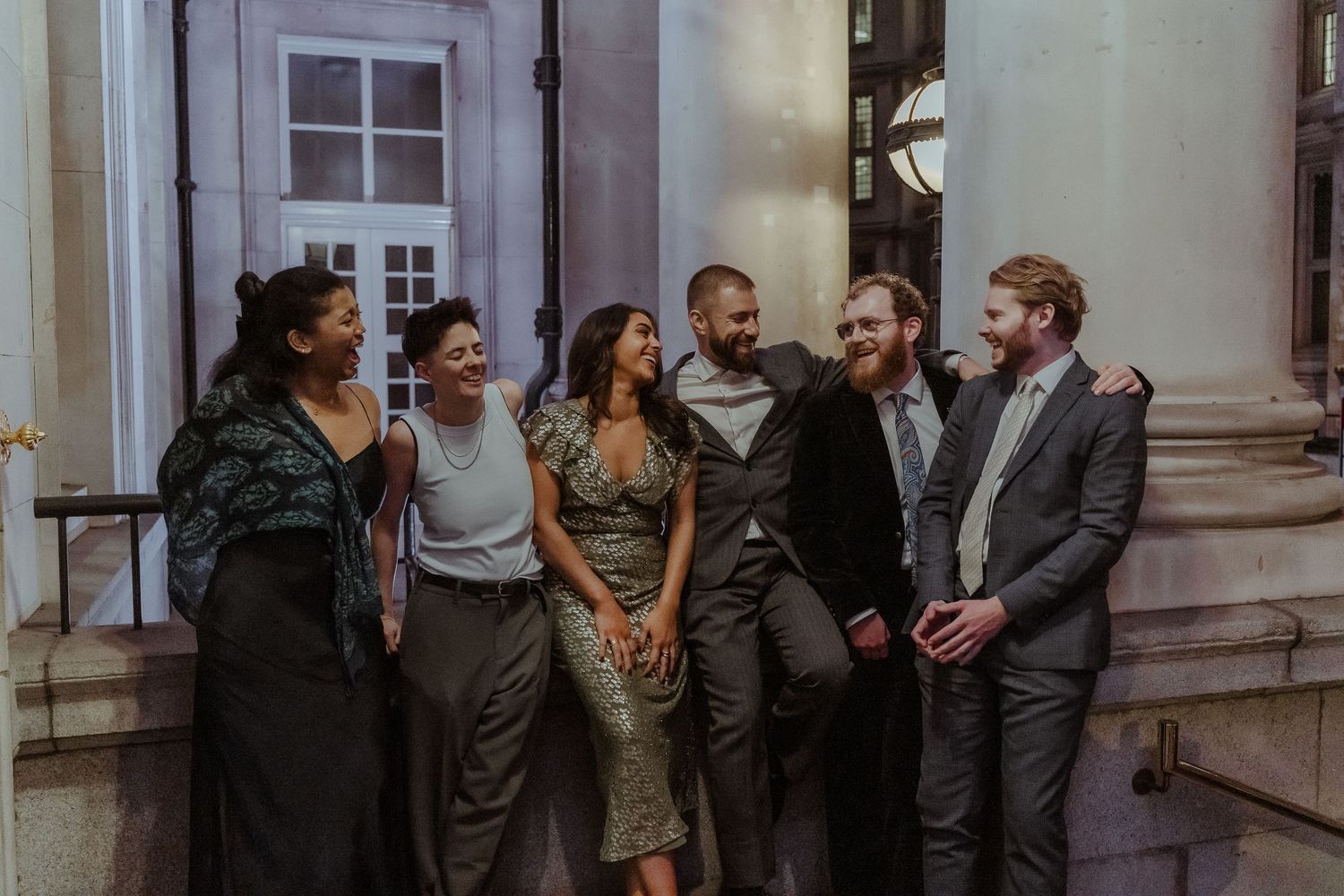 Six friends laughing and socializing outdoors at night near a classic building entrance.
