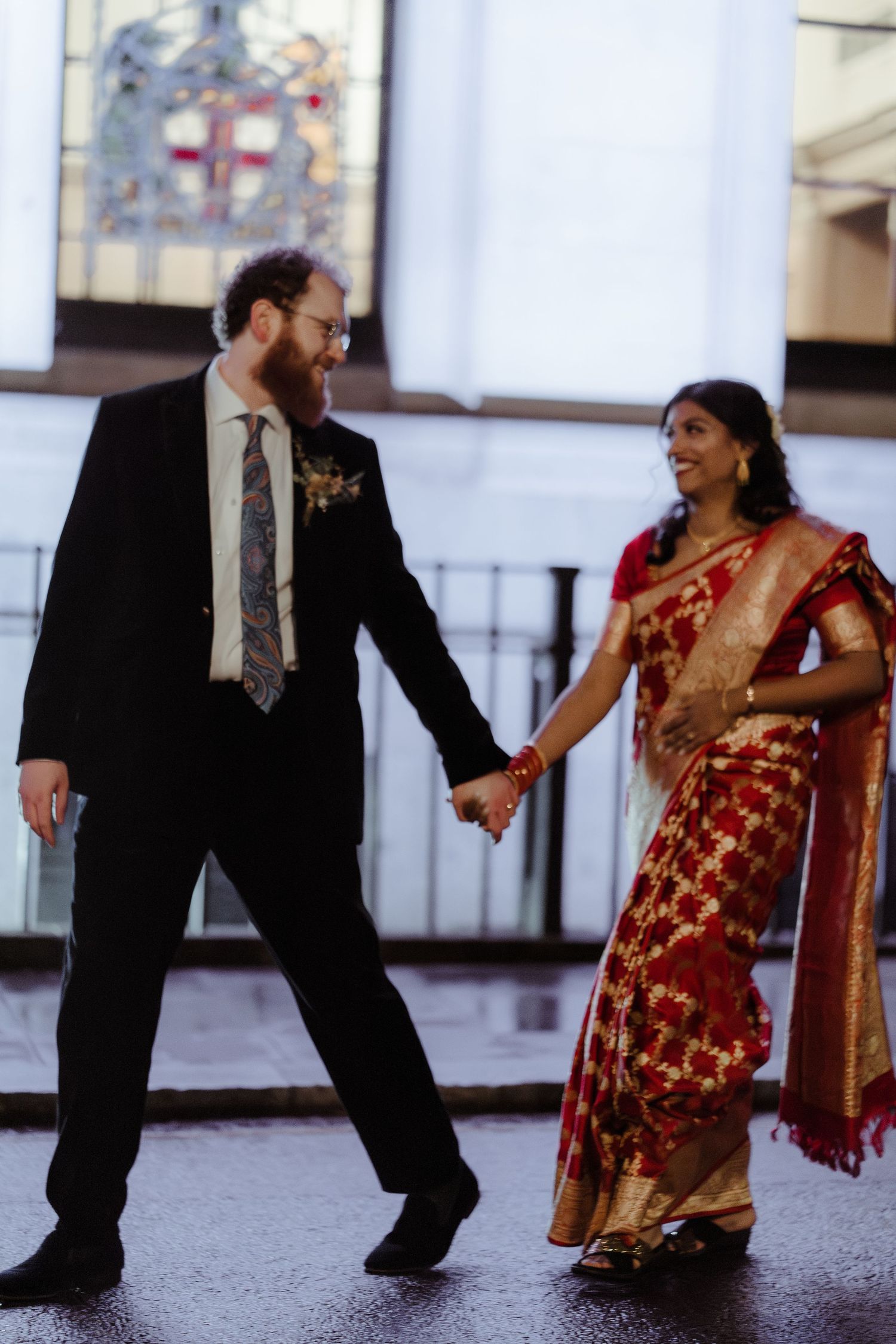 Couple holding hands outdoors, groom in black suit and bride in red gold saree, smiling at each other on their wedding day.