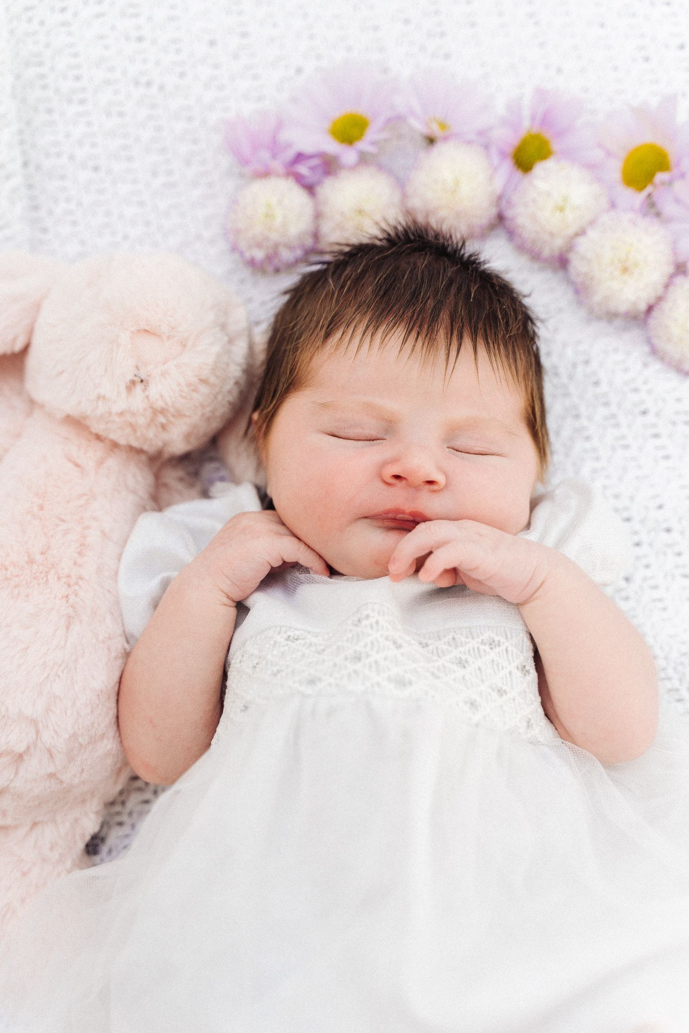 Sleeping newborn baby in white dress lying beside pink teddy bear and purple flowers on white blanket.