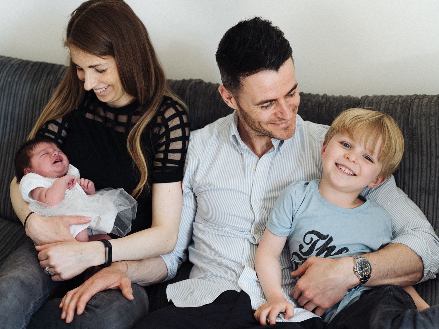 Happy family of four sitting on sofa, mother holding newborn baby, father and young blonde boy smiling together.