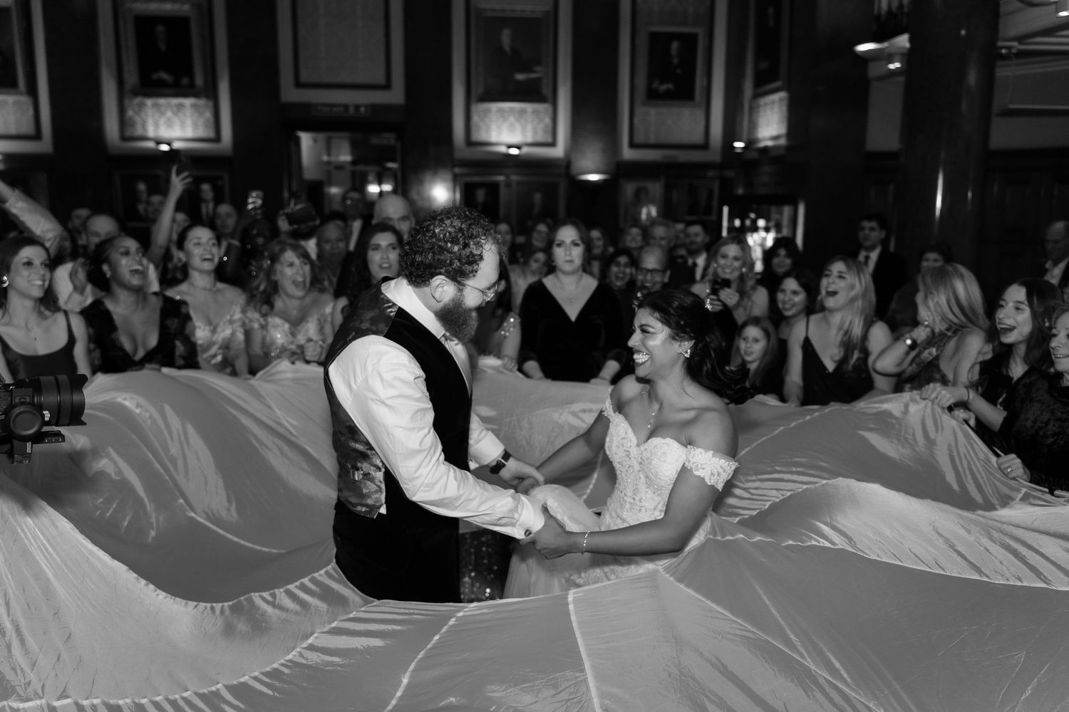 Black and white photo of bride and groom sharing first dance, surrounded by cheering guests at elegant wedding reception.