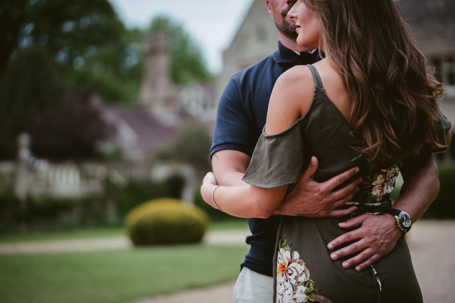 Couple embracing outdoors near a charming stone building, woman in olive dress hugging man in navy polo shirt.