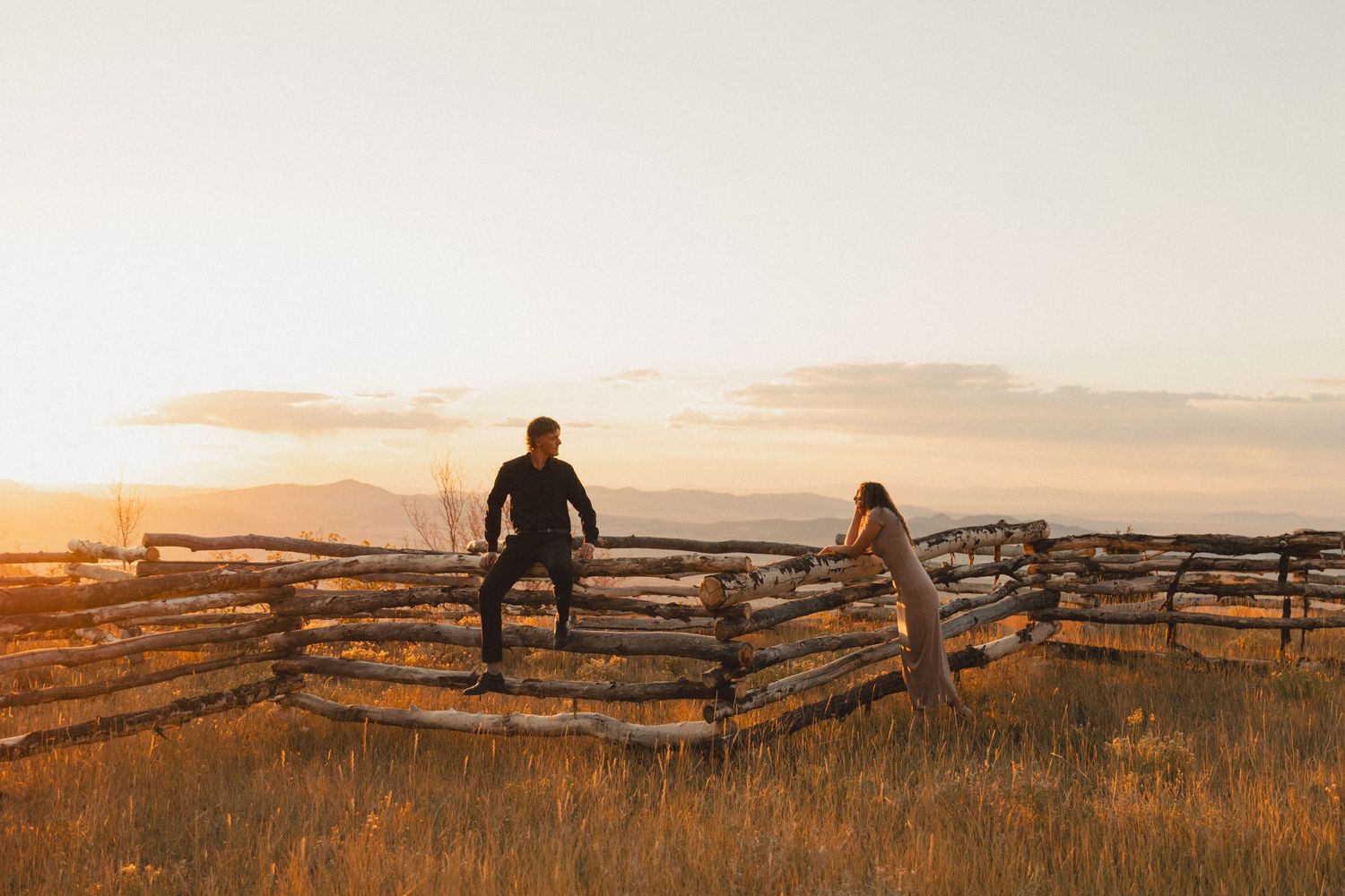 Man sitting on a wooden fence, gazing at his woman in a serene field, capturing a moment of connection.