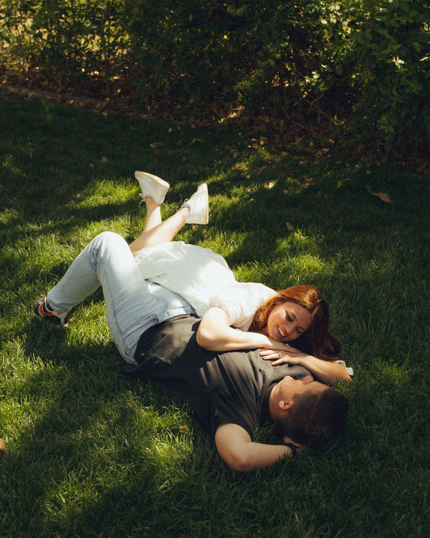 Couple laughing together while lying on the grass in a park, sharing a joyful and carefree moment.