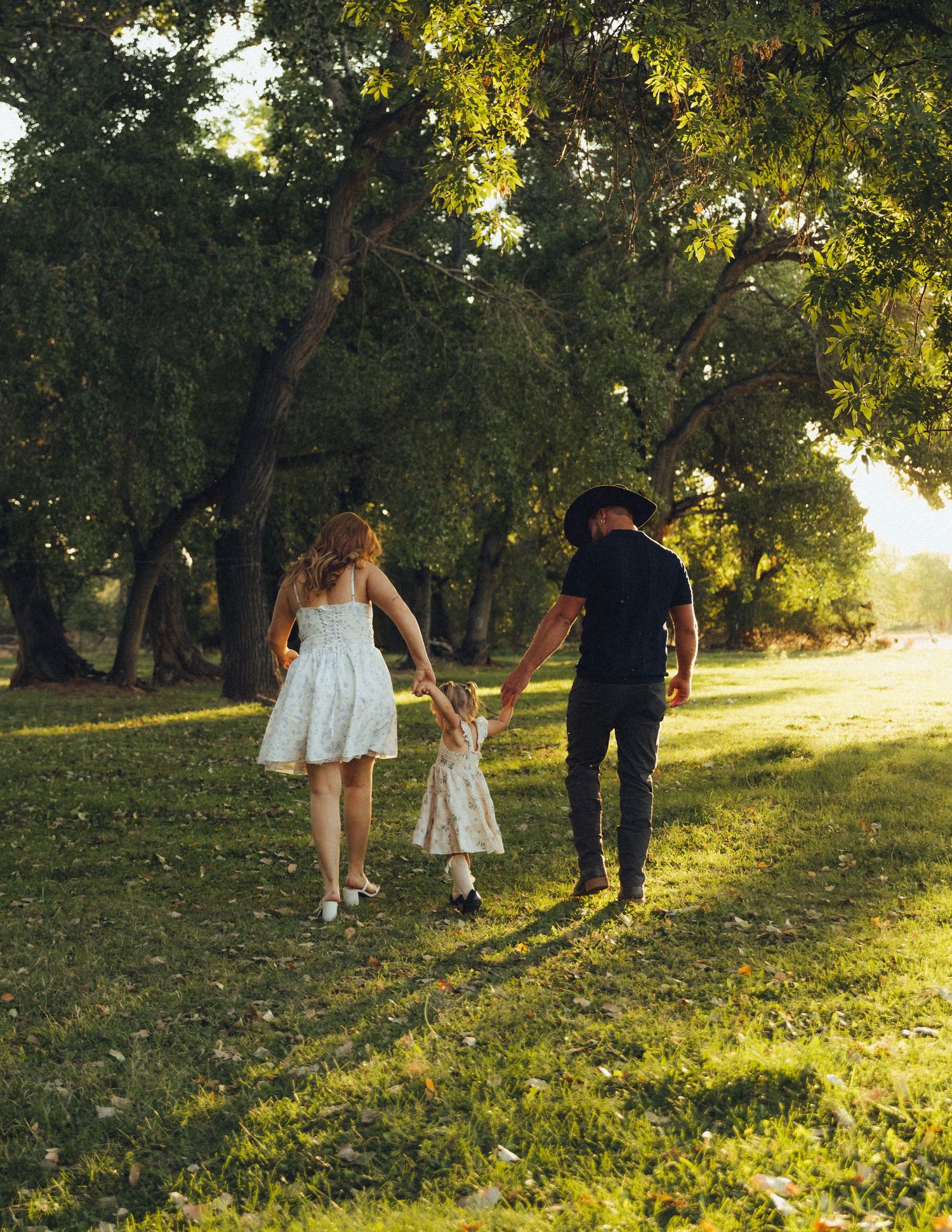 Family of three – woman, man, and little girl – holding hands and walking together into a lush, green setting.