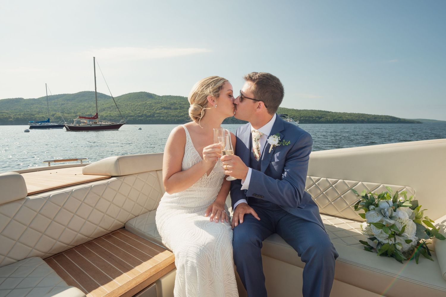 A young bride and groom embrace in a kiss while toasting champagne on a classic wooden boat during their wedding recepti