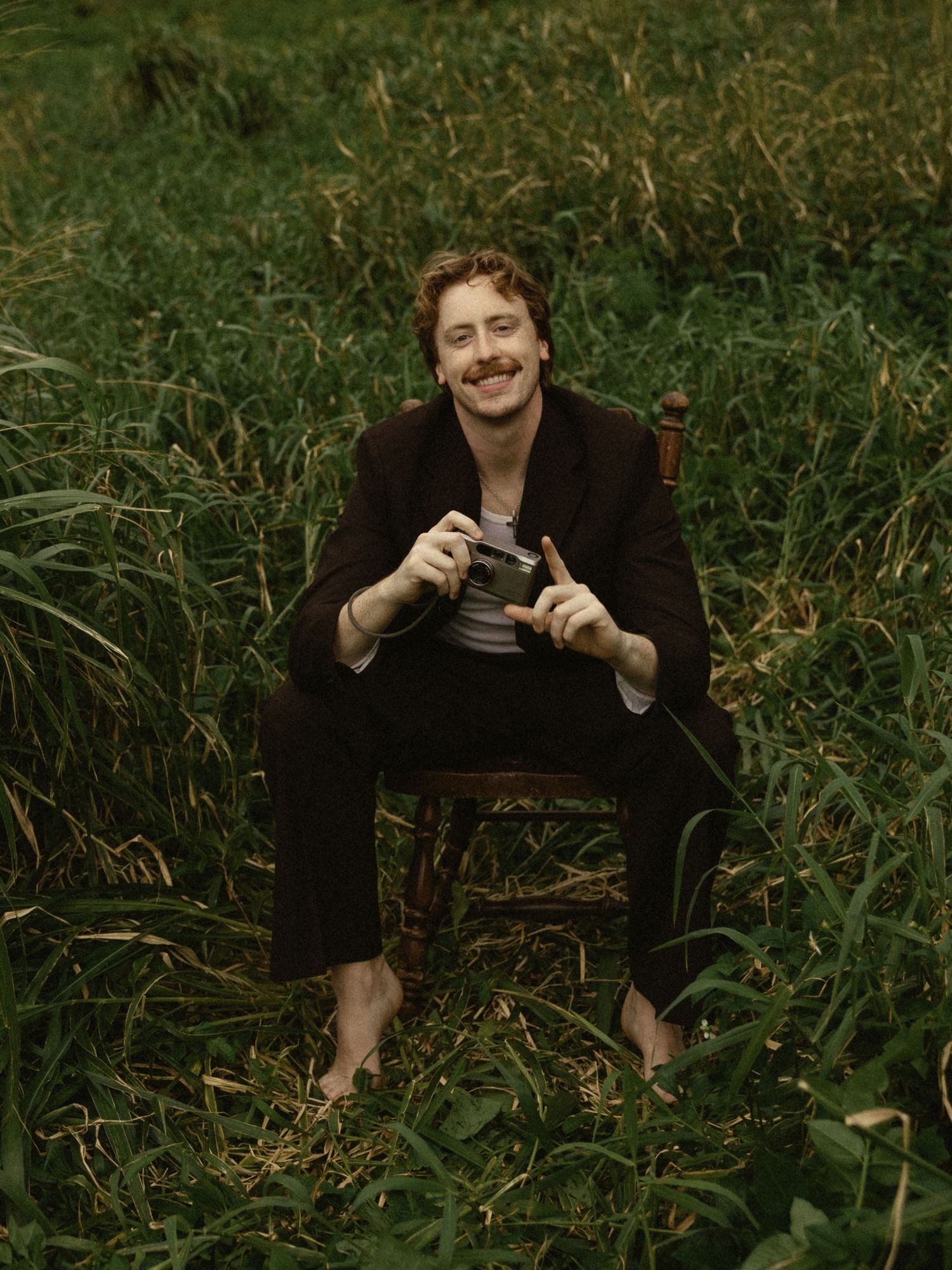 A photographer sits in tall grass holding a vintage camera in a series of candid outdoor shots.
