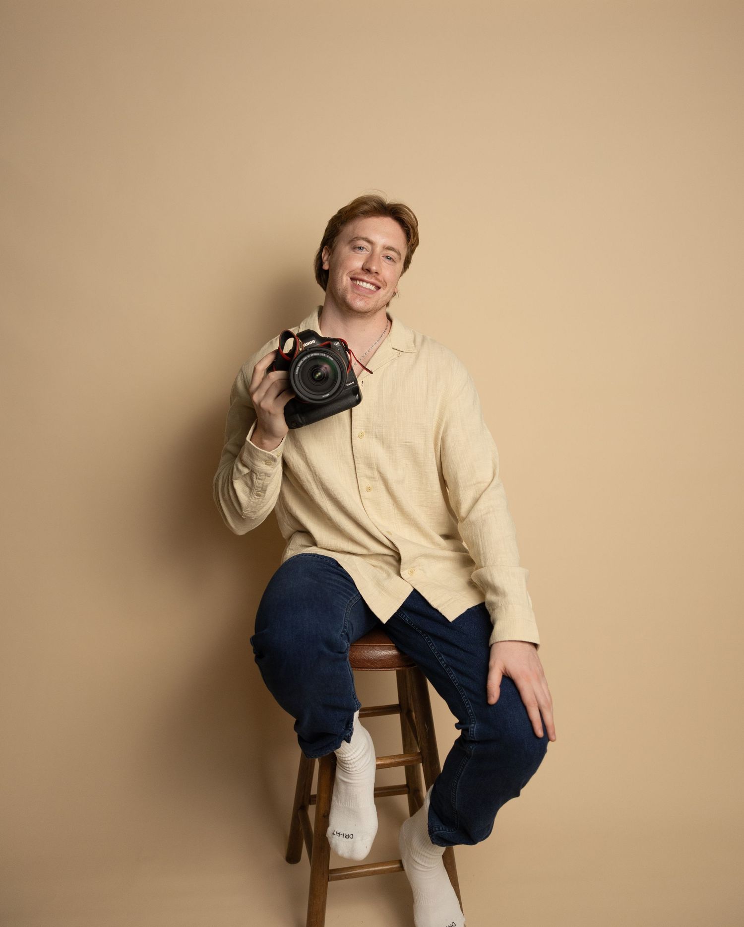 Creative portrait of photographer holding professional camera while seated on wooden stool.