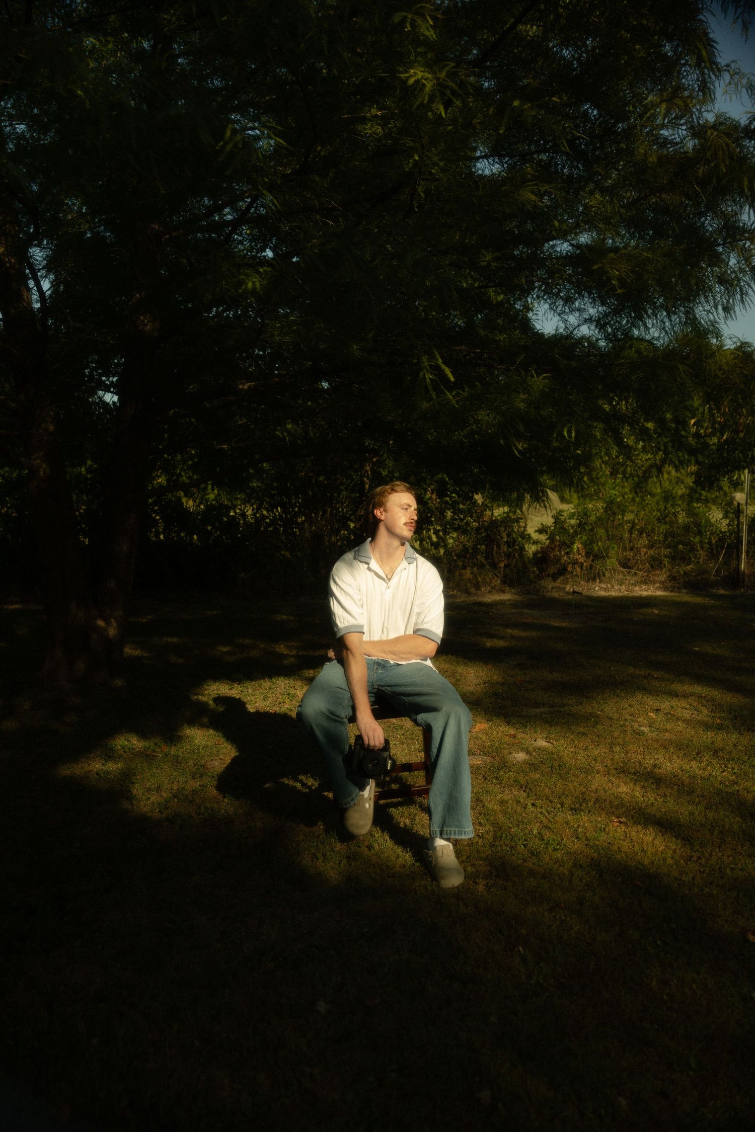 A figure sits on a wooden chair in a sun-dappled garden setting during golden hour.