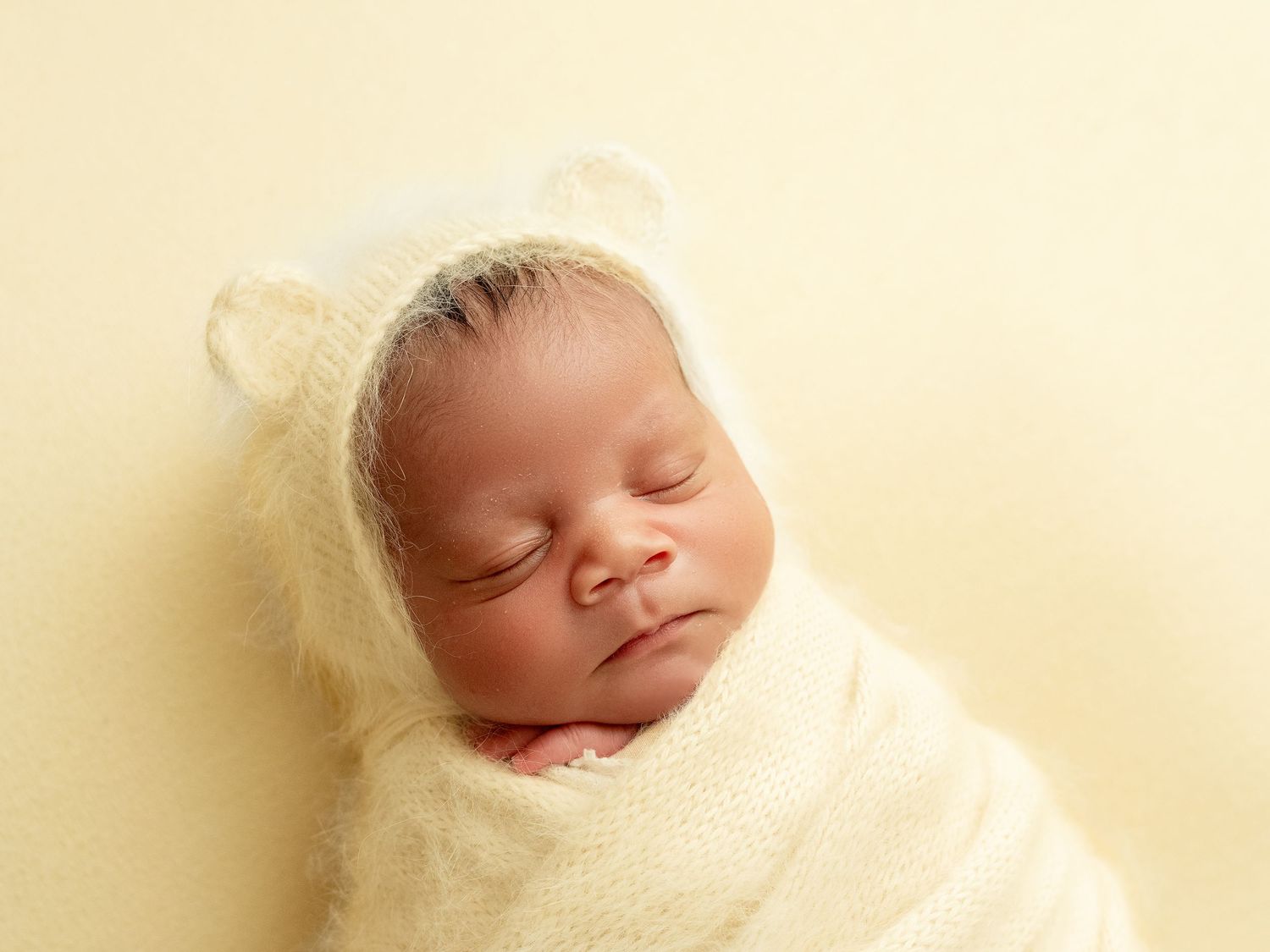 A sleeping newborn baby wrapped in a soft cream-colored blanket lies peacefully against a light background.