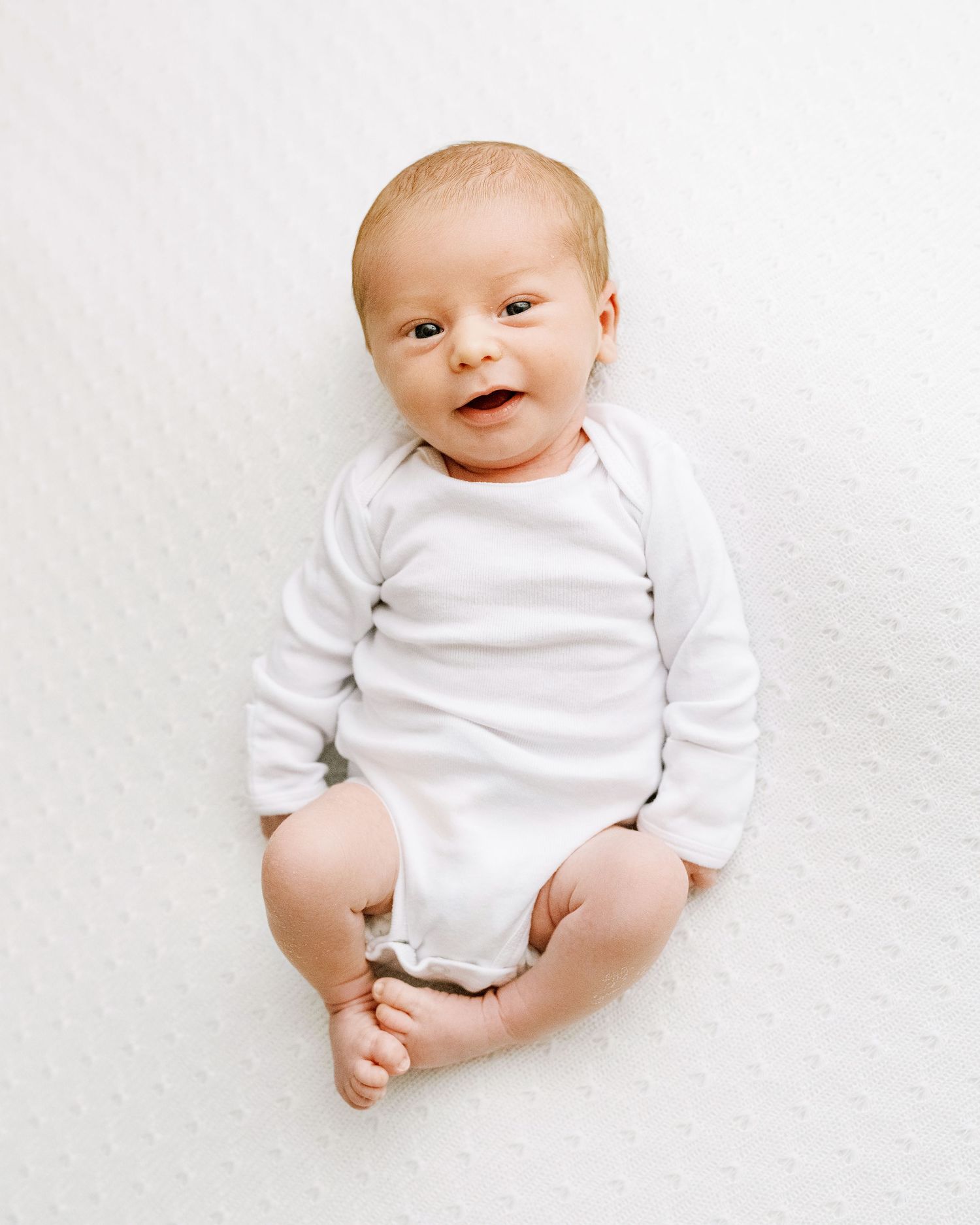 A baby dressed in a white long-sleeve onesie lies on a white background with a happy expression.
