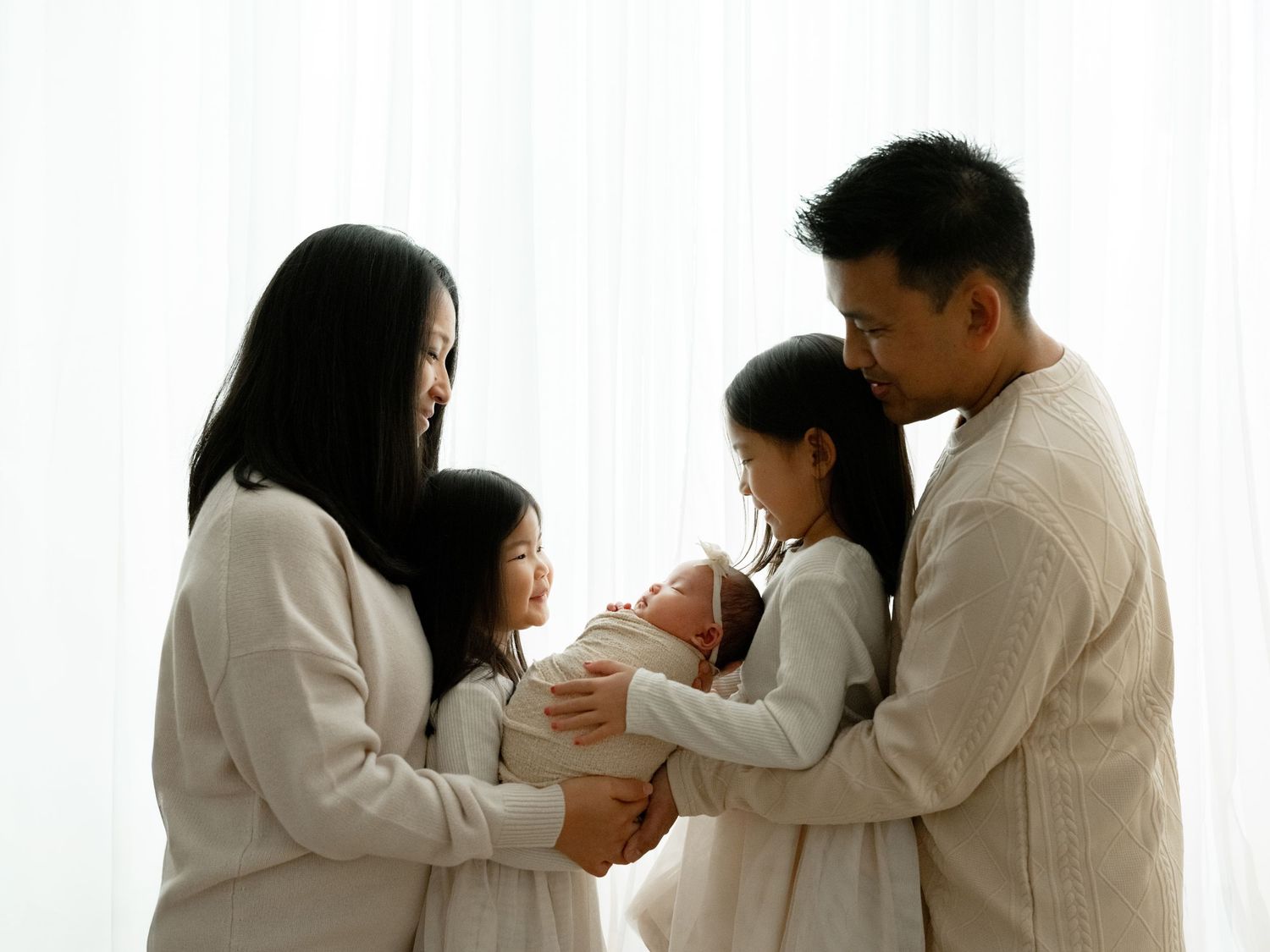 Asian family of four in white outfits admiring their newborn baby together in a bright, softly lit room.