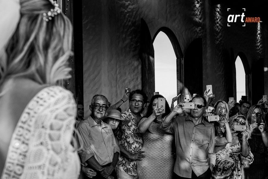 Black and white photo of wedding guests smiling and taking photos with smartphones in an elegant arched venue.