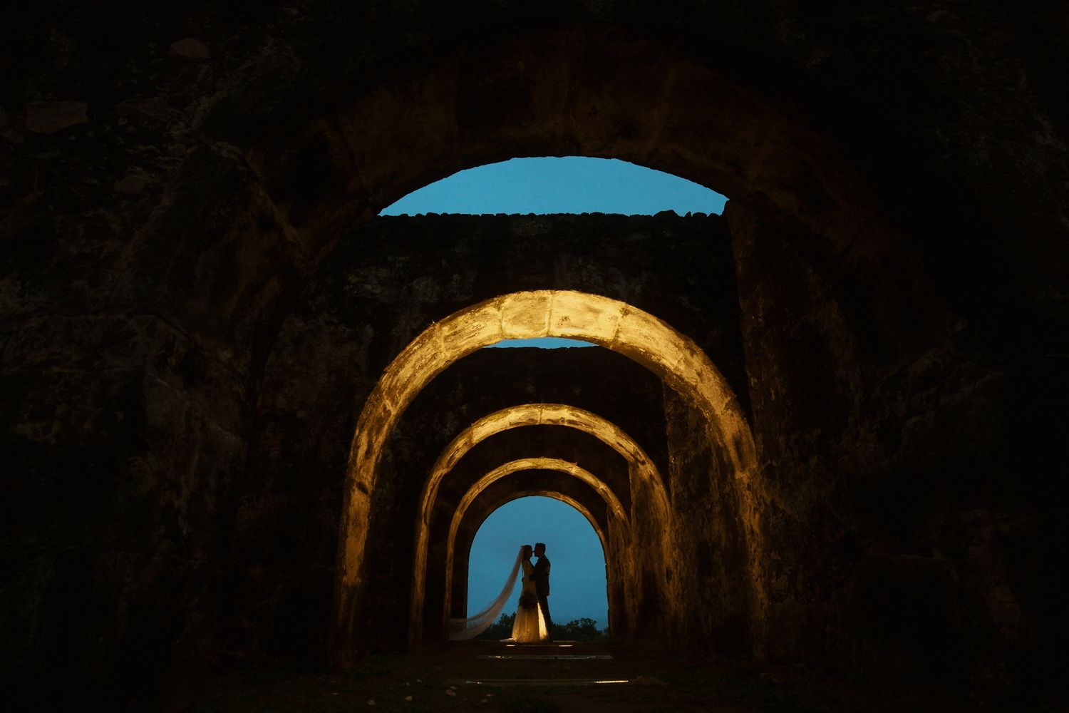 Wedding couple sharing a romantic kiss framed by dramatic stone archways at night.