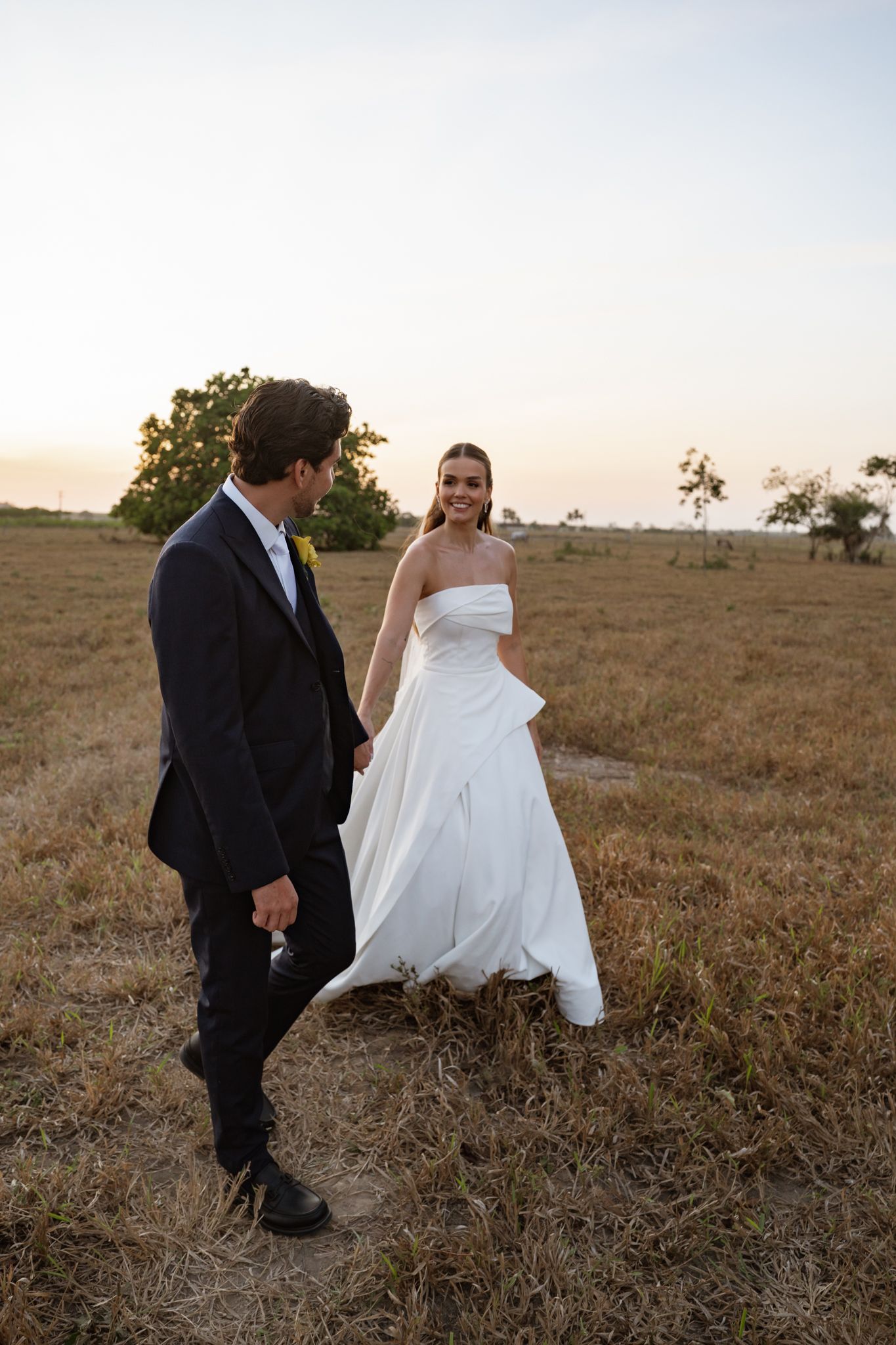 Bride and groom walking hand in hand through a golden field at sunset during their outdoor wedding.