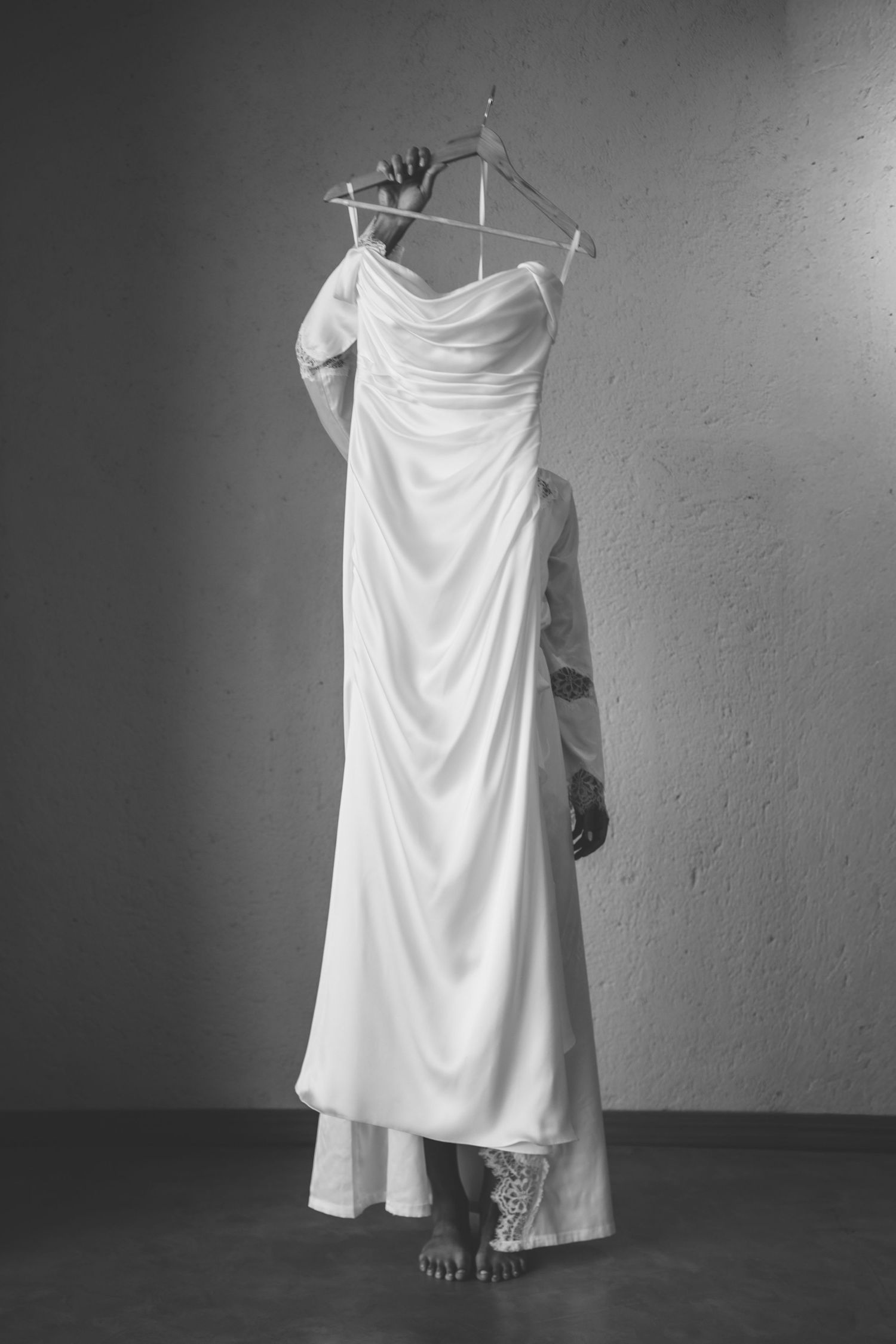 Monochrome image of a person holding a flowing strapless bridal gown on a hanger against a gray wall.