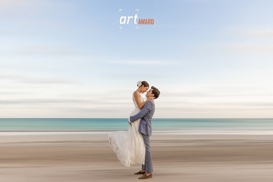 Couple sharing a romantic kiss on a sandy beach, groom lifting bride in wedding attire with turquoise ocean backdrop.