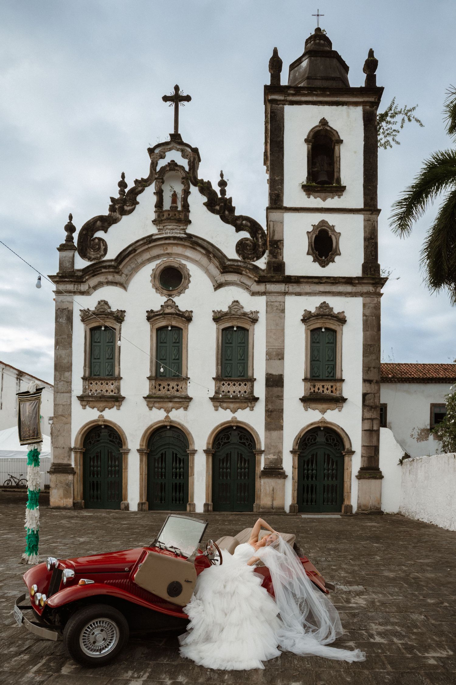 Bride in white gown posed on vintage red car before ornate Brazilian colonial church with bell tower.