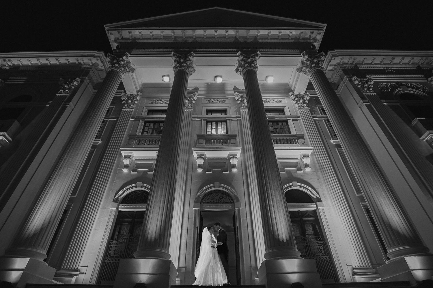 Black and white night photo of wedding couple embracing before a grand neoclassical building.