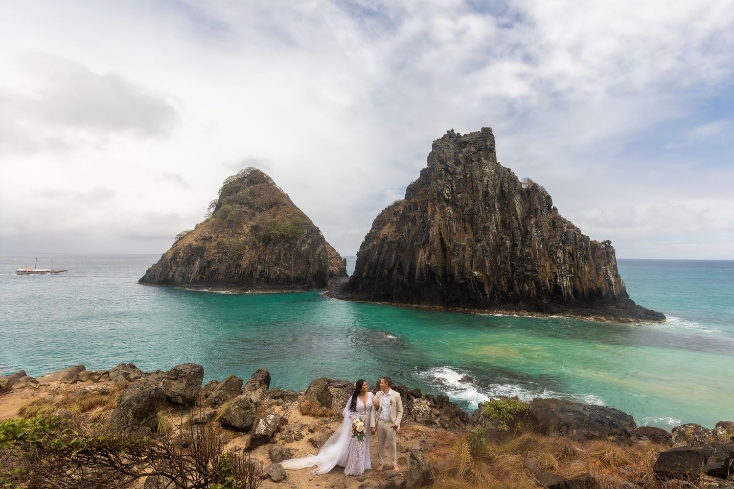 Couple in white wedding attire poses on rocky cliffs with turquoise ocean and dramatic twin rock formations in background.