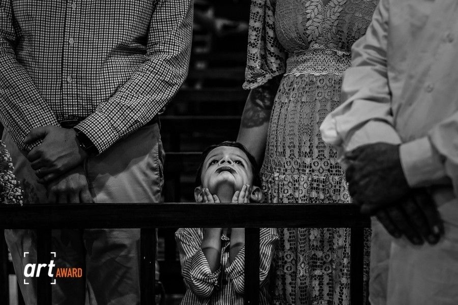 Black and white photo of a child peeking through a fence at a wedding ceremony.